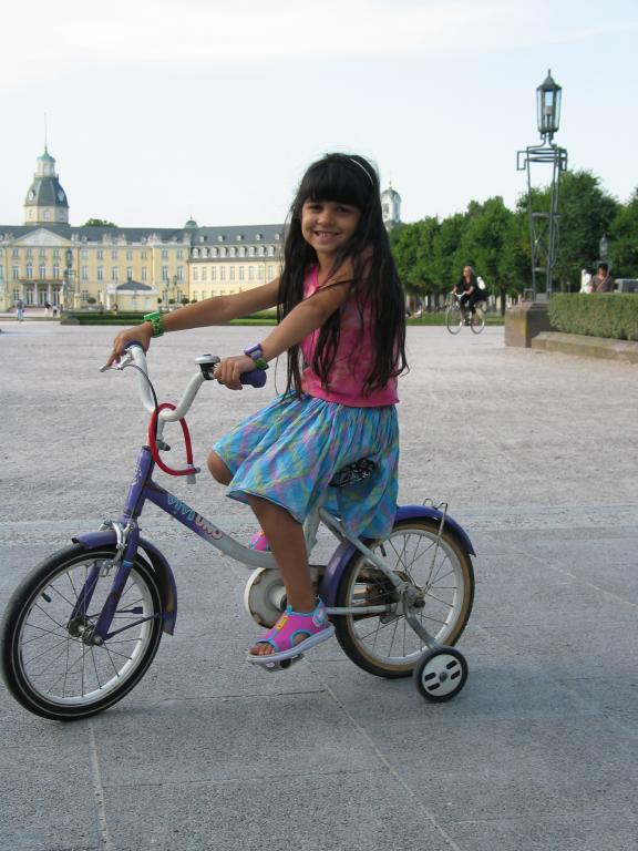 Xandie poses with her "new" bike.  When we arrived, the head of the
International Department offered to let us have the use of some
abandoned bicycles.  The first one we found for Xandie turned out to
be irreparable, but this one was actually better.  Note that it only
has one training wheel.  It took no time at all for Xandie to learn
that all good cyclists lean to the left!